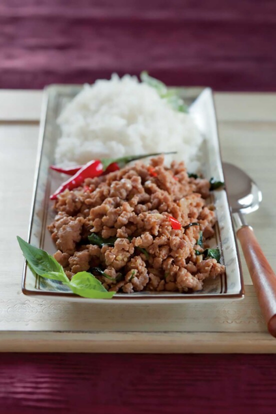 A wooden cutting board with a rectangular plate and spoon on top, the plate has a mound of rice on the back half. On the front half, there's a pile of ground pork, basil, and peppers.