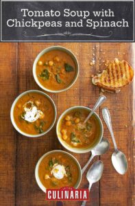 Three white bowls on a wooden table with soup spoons and half a grilled cheese sandwich. Each bowl is filled with tomato soup with chickpeas and spinach and topped with yogurt.