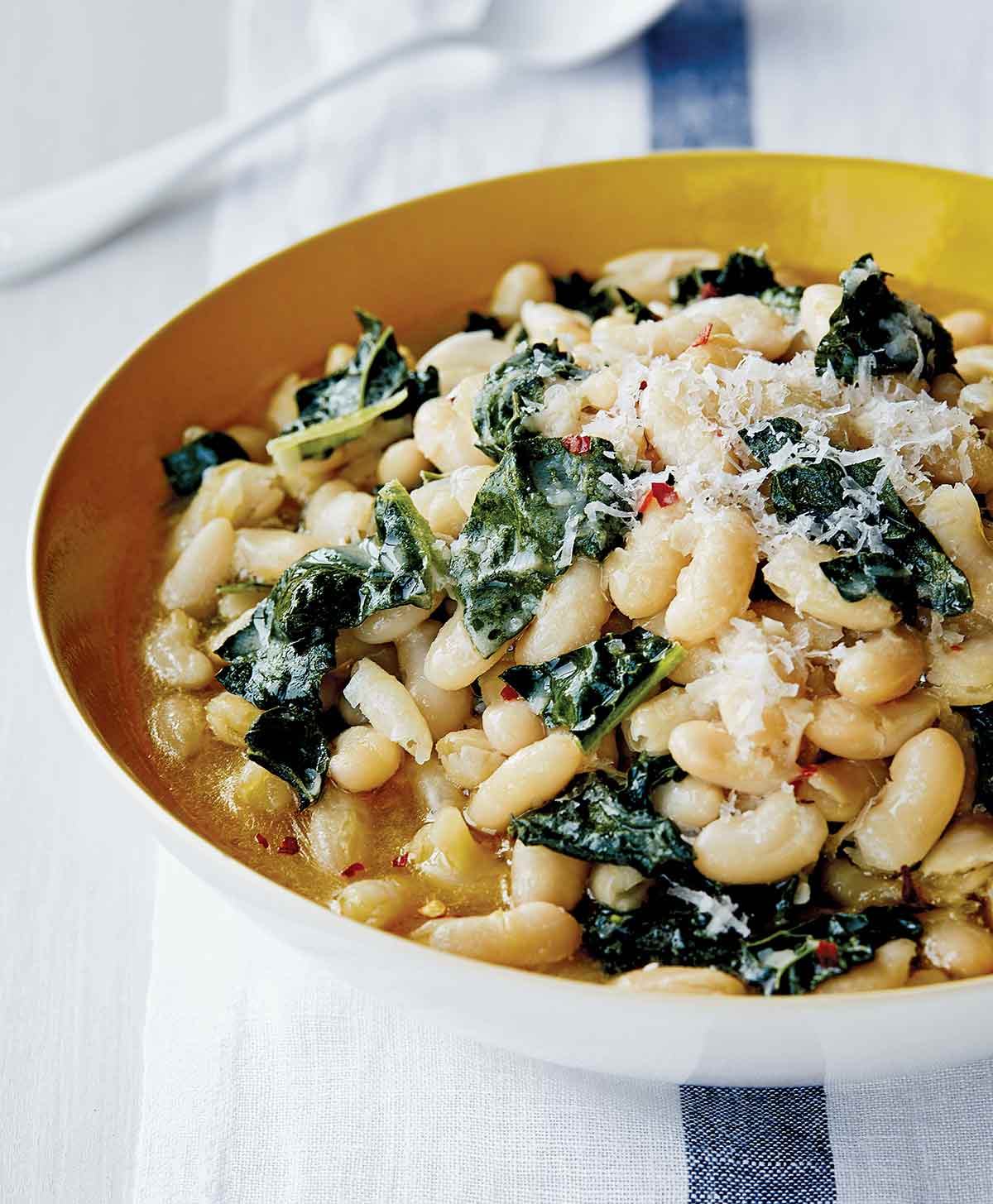 A bowl of white beans and Tuscan kale, sprinkled with grated Parmesan cheese, with a spoon.