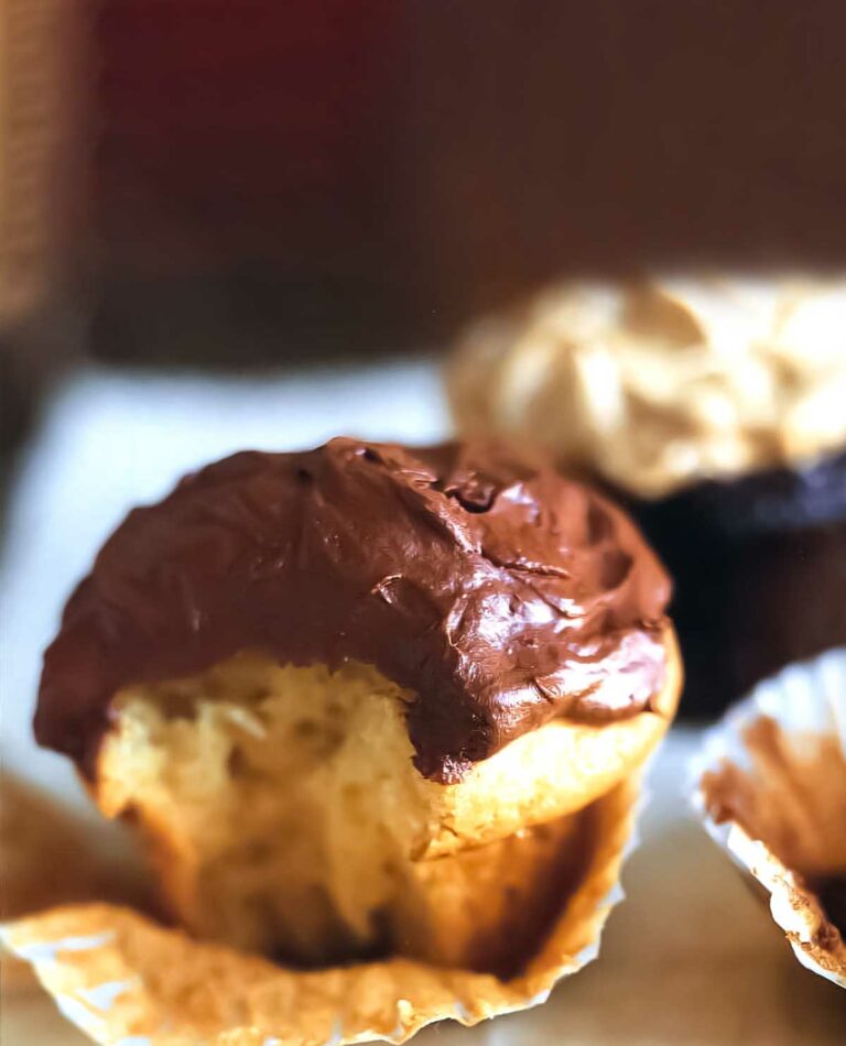A white cupcake with chocolate frosting, with the paper peeled off and a bite taken out of it.