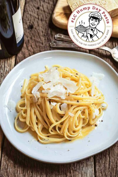 A white plate on a wooden table, filled with white wine linguine covered with black pepper and shavings of Parmesan, with a chunk of Parmesan and a bottle of white wine next to it.