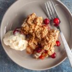 A plate with a serving of apple cranberry crisp, a scoop of vanilla ice cram, and a fork on the side.