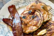 Three blueberry buttermilk pancakes and two slices of bacon, all drizzled with maple syrup, on a decorative plate with a fork resting beside the food.