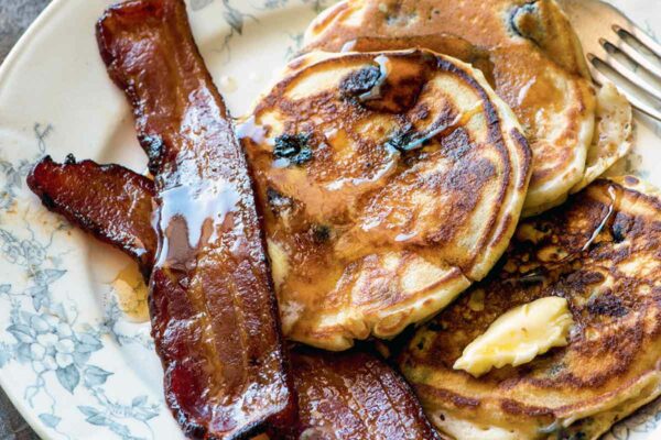 Three blueberry buttermilk pancakes and two slices of bacon, all drizzled with maple syrup, on a decorative plate with a fork resting beside the food.