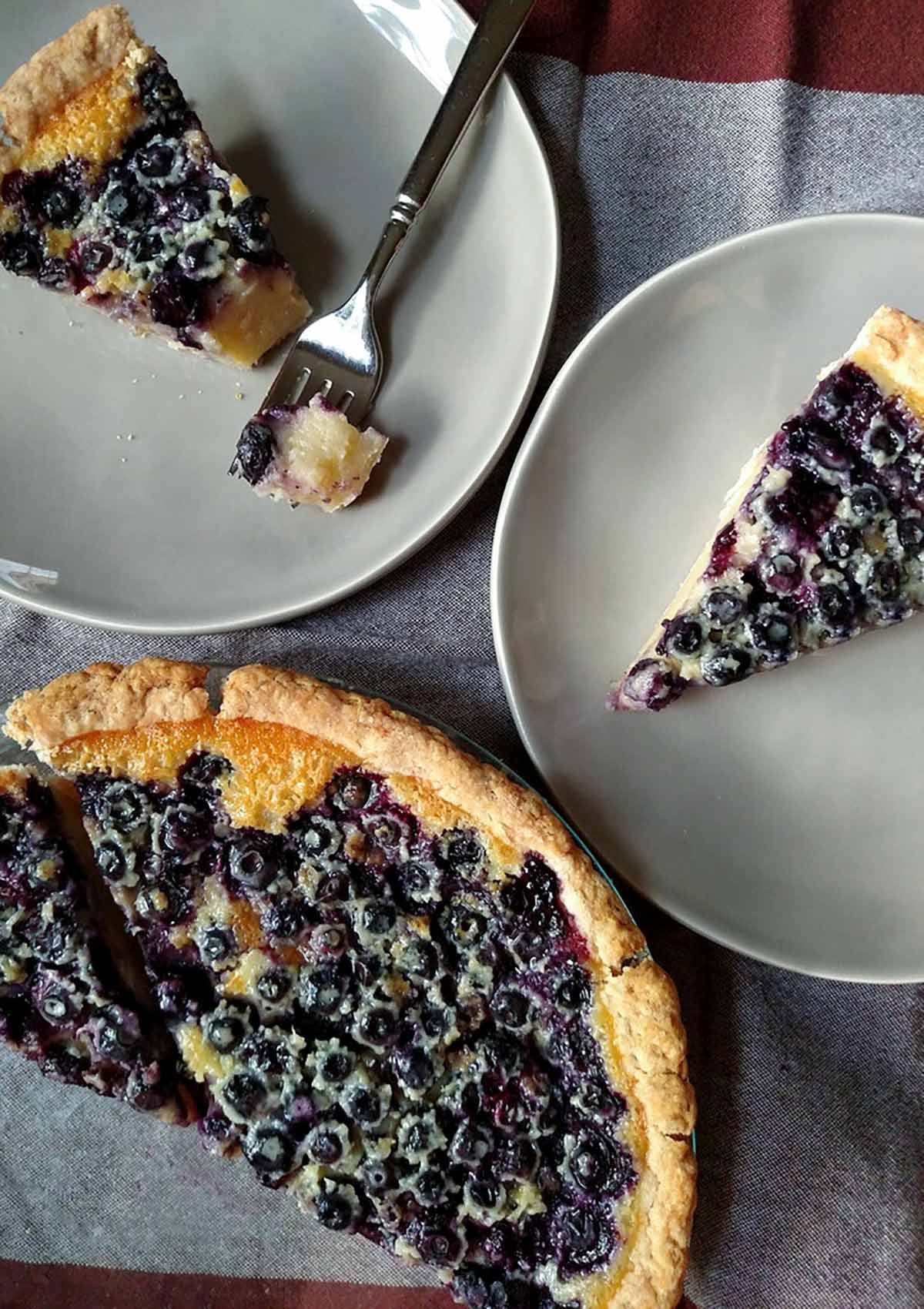 A blueberry custard pie in a pie plate, with 2 dessert plates with a slice of pie on them, one with a fork and a bite missing.
