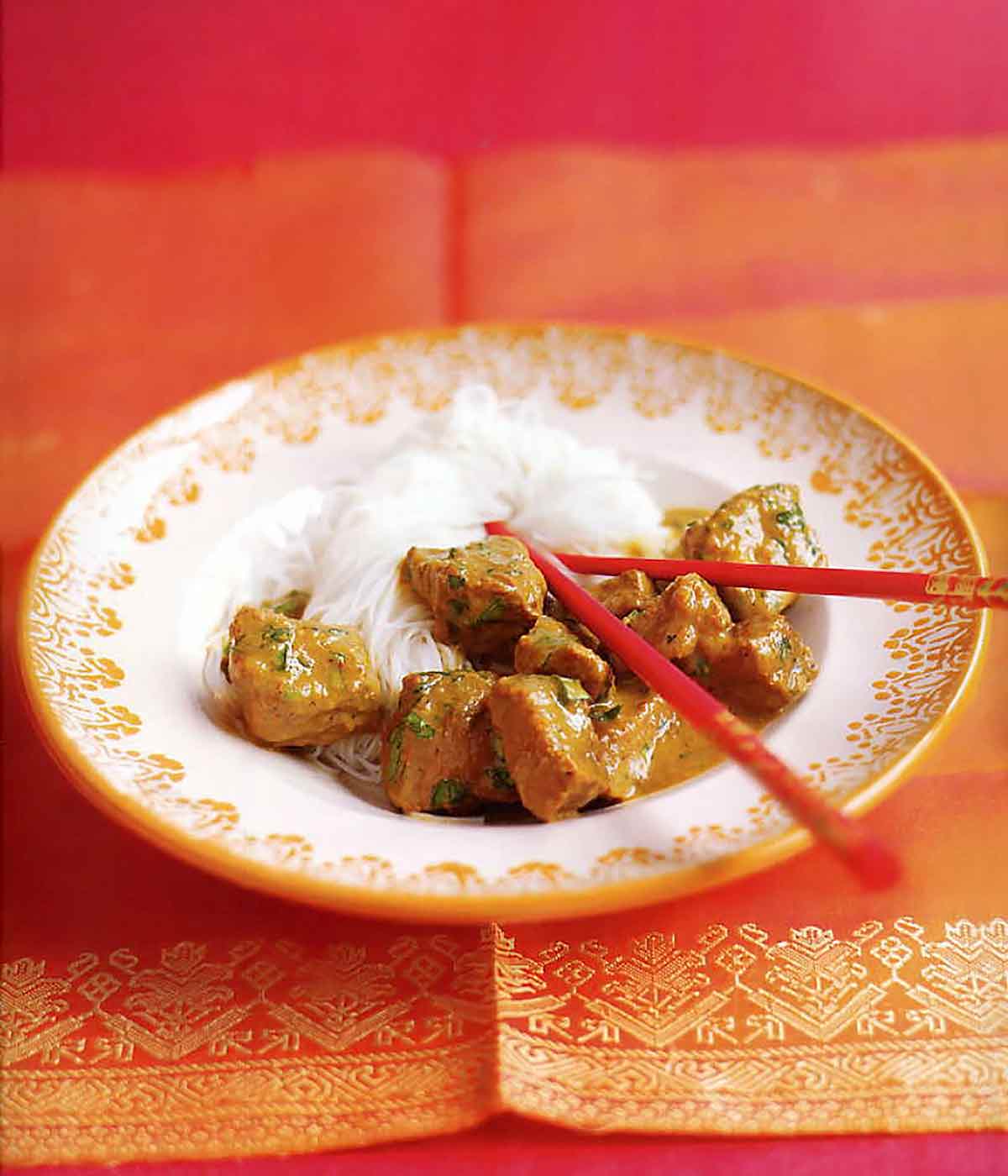 An orange tablecloth with a white and gold pattern filled with rice vermicelli, Burmese beef curry and red chopsticks.