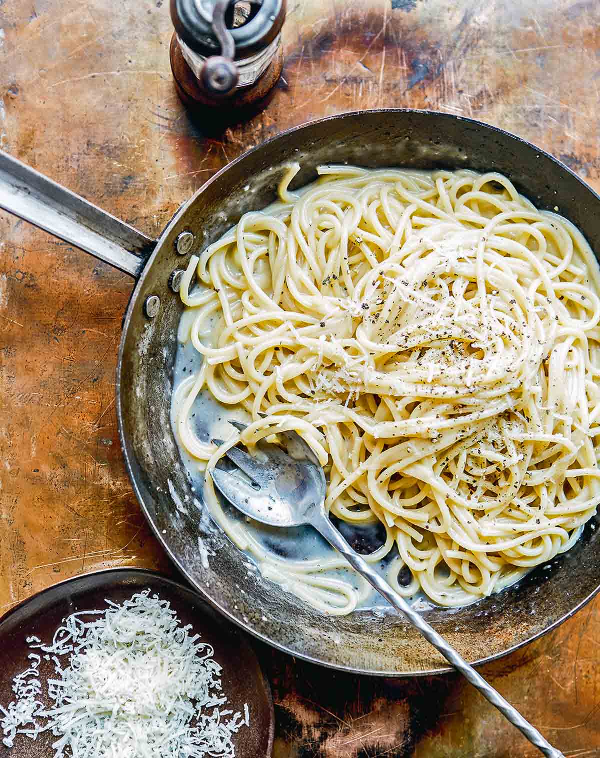 En metallpanna fylld med krämig pasta täckt med strimlad parmesan och hackad peppar, med en stor serveringssked.  En skål med parmesan och en pepparkvarn sitter bredvid.