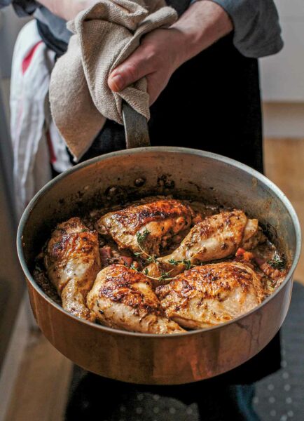 Chicken with mustard in a large metal skillet garnished with thyme, being held by a man.