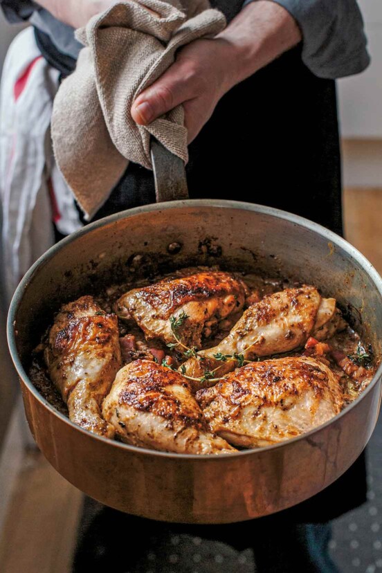 Chicken with mustard in a large metal skillet garnished with thyme, being held by a man.