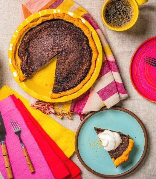 A table with colourful napkins, a plate with a slice of pie , a cup of coffee, and a yellow pie plate with a chocolate buttermilk pie that's missing one slice.