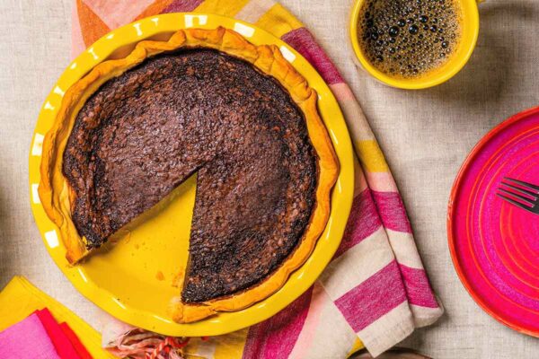 A table with colourful napkins, a plate with a slice of pie, a cup of coffee, and a yellow pie plate with a chocolate buttermilk pie that's missing one slice.