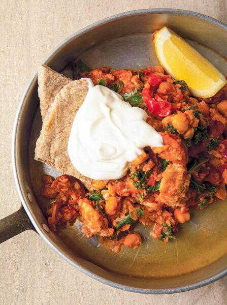 A saucepan filled with a serving of cod and chorizo stew, 2 pita quarters, a dollop of sour cream, and a lemon wedge.