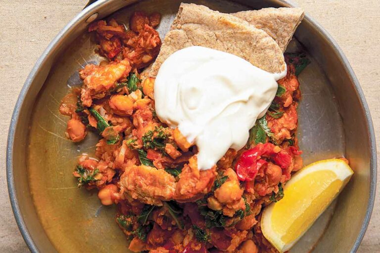 A saucepan filled with a serving of cod and chorizo stew, 2 pita quarters, a dollop of sour cream, and a lemon wedge.