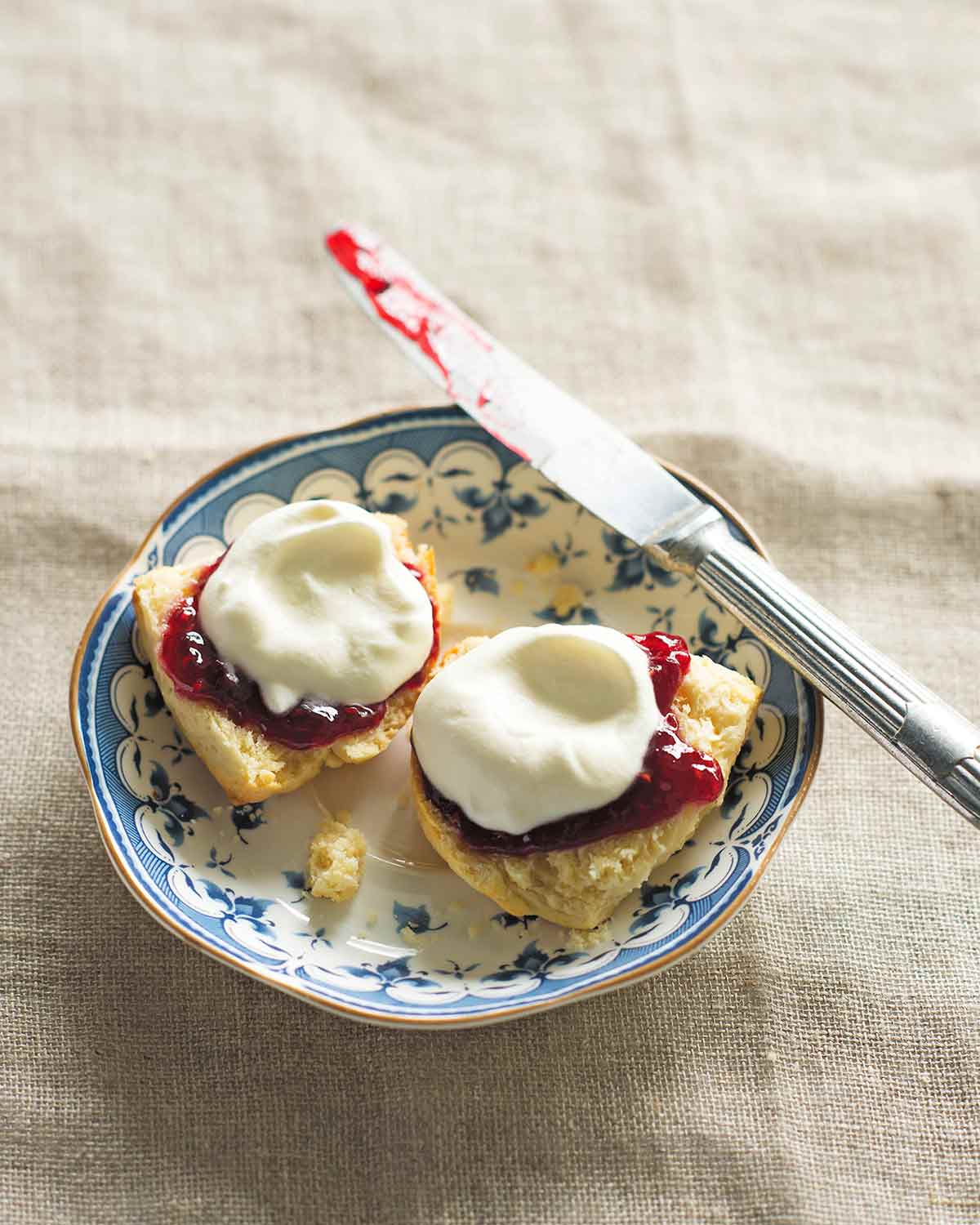 A blue and white saucer with a knife and 2 halves of a cream scone, topped with jam and whipped cream.