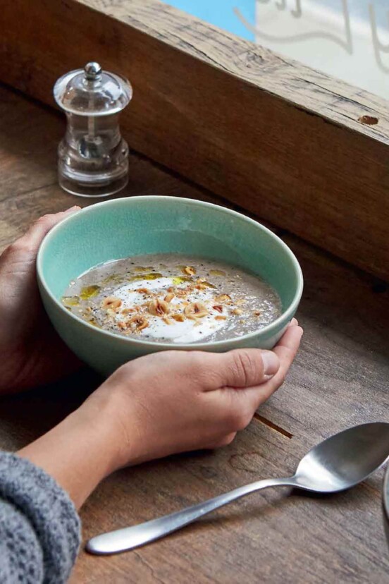 2 hands holding a pale green bowl filled with creamy mushroom soup, topped with olive oil yogurt, and hazelnuts. Salt and pepper shakers, and a spoon sit next to it.