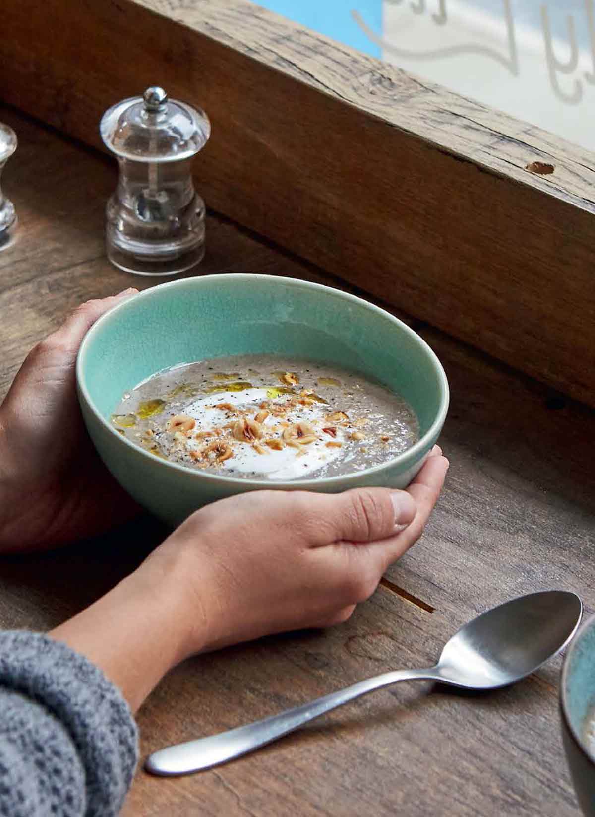2 hands holding aloft green bowl filled with creamy mushroom soup, topped with olive oil yogurt, and hazelnuts. Salt and pepper shakers, and a spoon sit next to it.