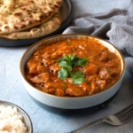 A bowl of quick butter chicken topped with cilantro, beside a fork, a bowl of rice, and naan bread.
