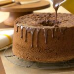 A Chinese five-spice chocolate chiffon cake on a wire rack, being drizzled with chocolate glaze from a yellow pitcher.