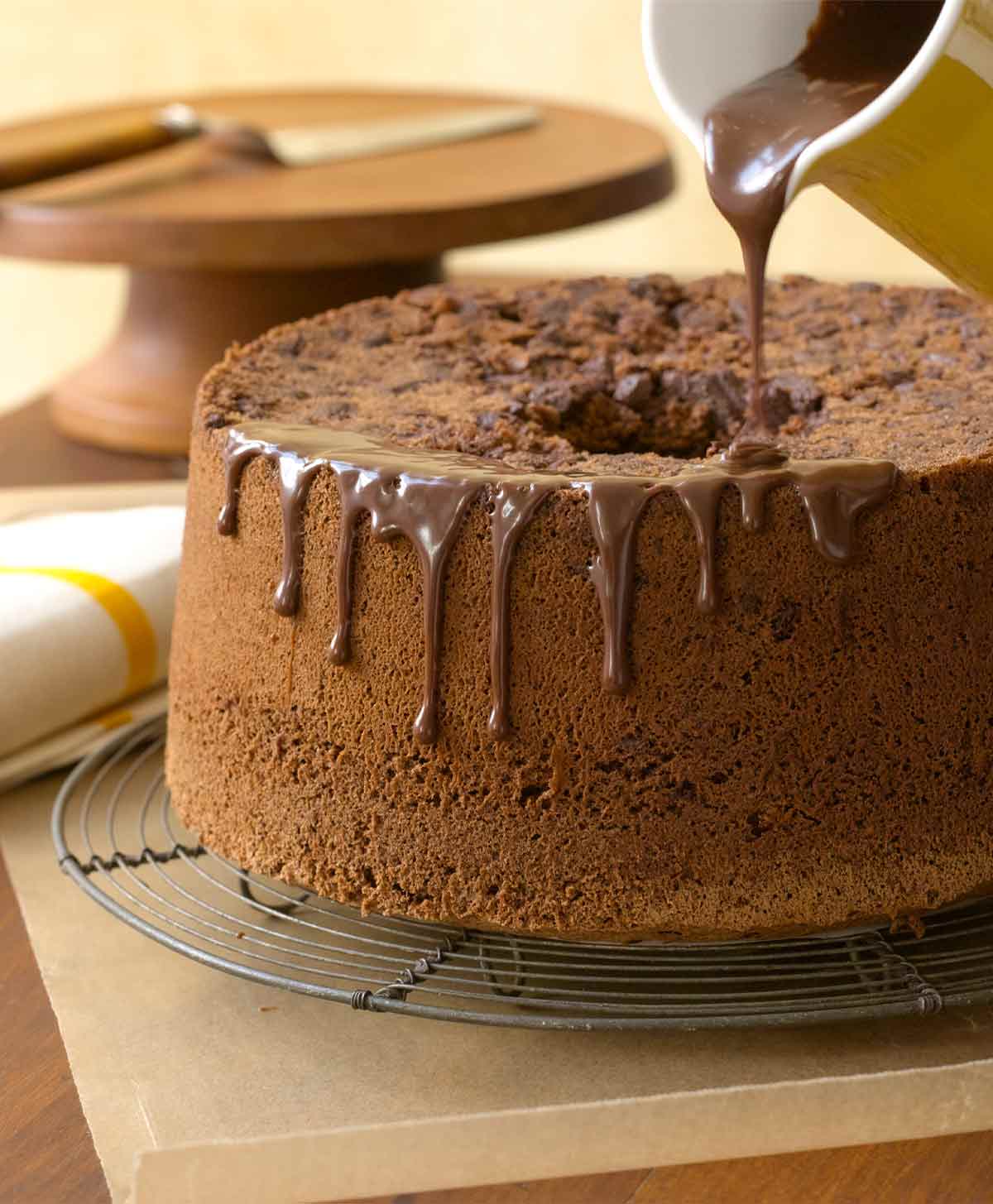 A Chinese five-spice chocolate chiffon cake on a wire rack, being drizzled with chocolate glaze from a yellow pitcher.
