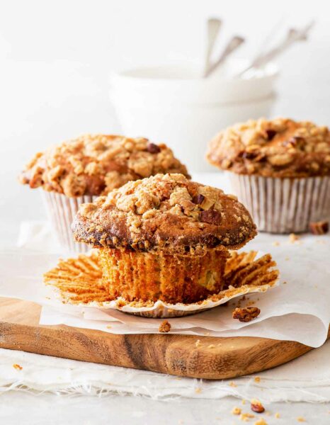 Three gluten-free apple pie muffins on a wooden chopping blocking with parchment paper and white bowls in the background.