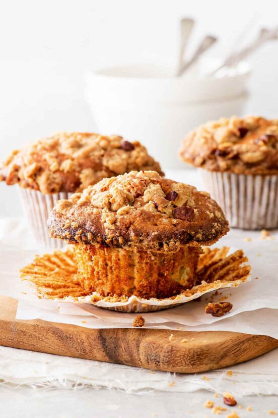 Three gluten-free apple pie muffins on a wooden chopping blocking with parchment paper and white bowls in the background.