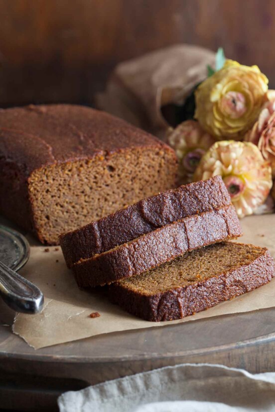 A loaf of gluten-free pumpkin bread lying on a table with 3 slices, with a butter knife and flowers in the background.