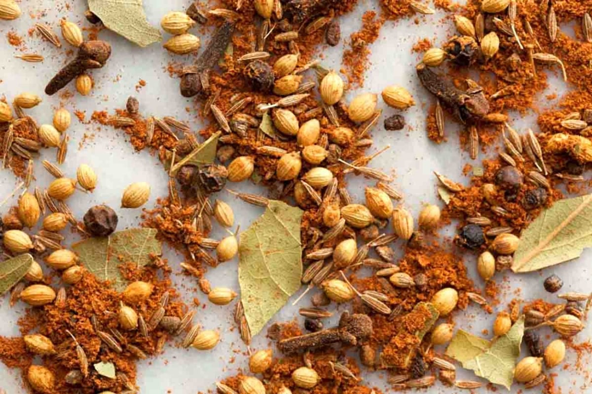 An overhead view of a vibrant blend of whole and ground Indian spices, including coriander seeds, bay leaves, cloves, and cumin, scattered on a light background.