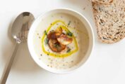 A white plate with a soup spoon and 2 slices of grainy bread, with a small white bowl filled with Jerusalem artichoke soup garnished with thyme, truffle oil, and Jerusalem artichoke chips.