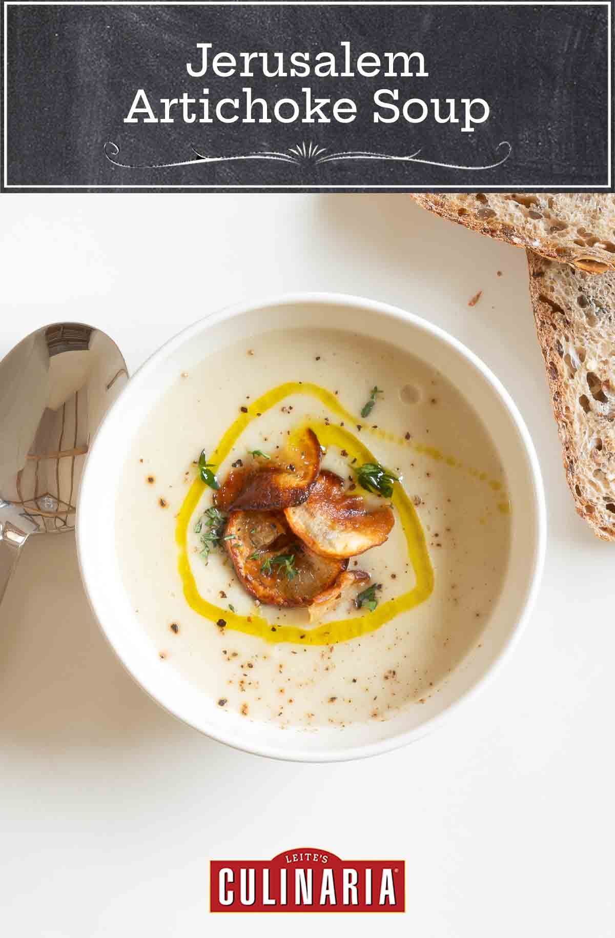 A white plate with a soup spoon and 2 slices of grainy bread, with a small white bowl filled with Jerusalem artichoke soup garnished with thyme, truffle oil, and Jerusalem artichoke chips.