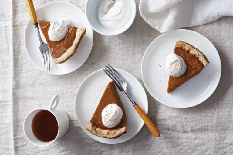 Maple pumpkin pie slices on 3 plates, topped with whipping cream. Flanked by a napkin, a cup of coffee, and a bowl of whipping cream.