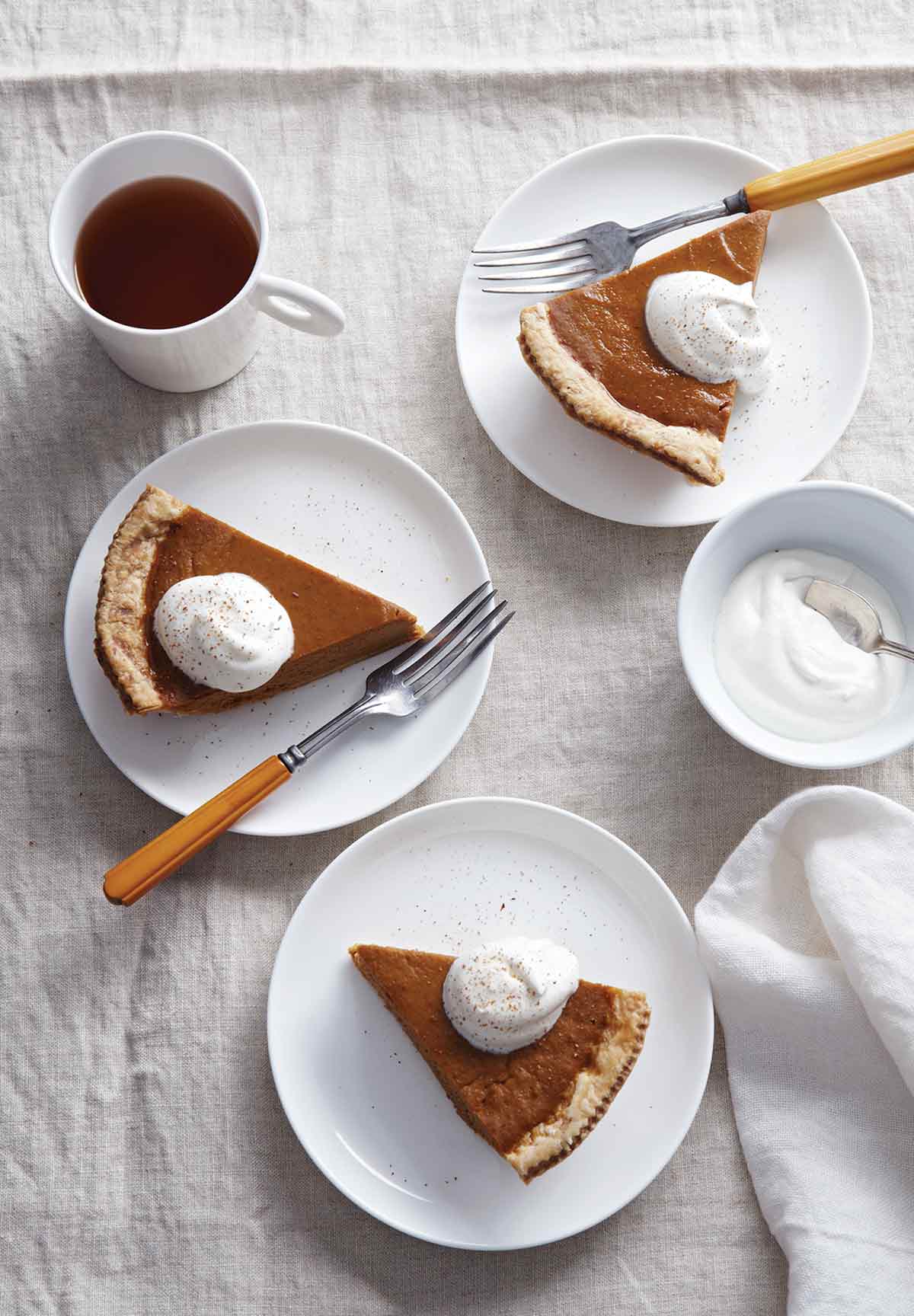 Maple pumpkin pie slices on 3 plates, topped with whipping cream. Flanked by a napkin, a cup of coffee, and a bowl of whipping cream.