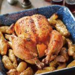 A blue enamel roast pan with a maple roast chicken, trussed and stuffed with lemon halves, sitting on a bed of fingerling potatoes. Glasses of red wine and salt and pepper in the background.