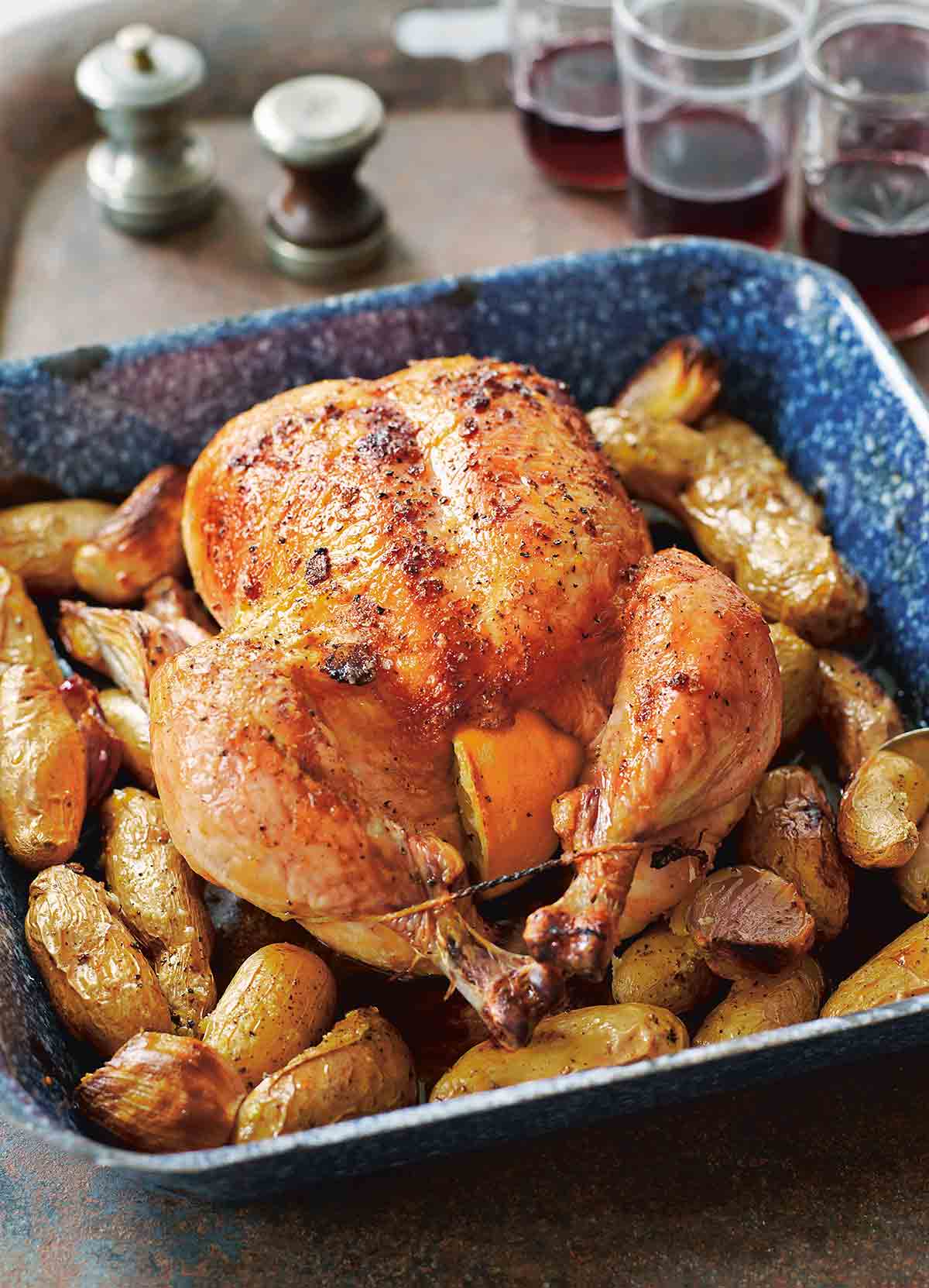A blue enamel roast pan with a maple roast chicken, trussed and stuffed with lemon halves, sitting on a bed of fingerling potatoes. Glasses of red wine and salt and pepper in the background.