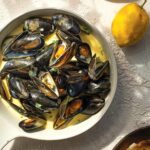 A white saucepan filled with mussels in a creamy white wine garlic sauce with lemons.
