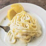 A white plate with a swirl of One-Pan Lemon Parmesan Linguine, with creamy sauce, a fork and 2 slices of lemon.