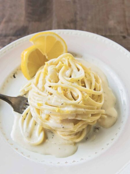 A white plate with a swirl of One-Pan Lemon Parmesan Linguine, with creamy sauce, a fork and 2 slices of lemon.