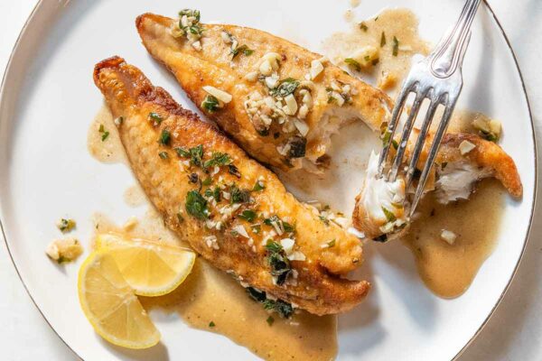 A white dinner plate with 2 filets of pan-fried bass with lemon garlic herb sauce, a fork, and 2 slices of lemon.