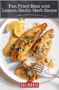 A white dinner plate with 2 filets of pan-fried bass with lemon garlic herb sauce, a fork, and 2 slices of lemon.