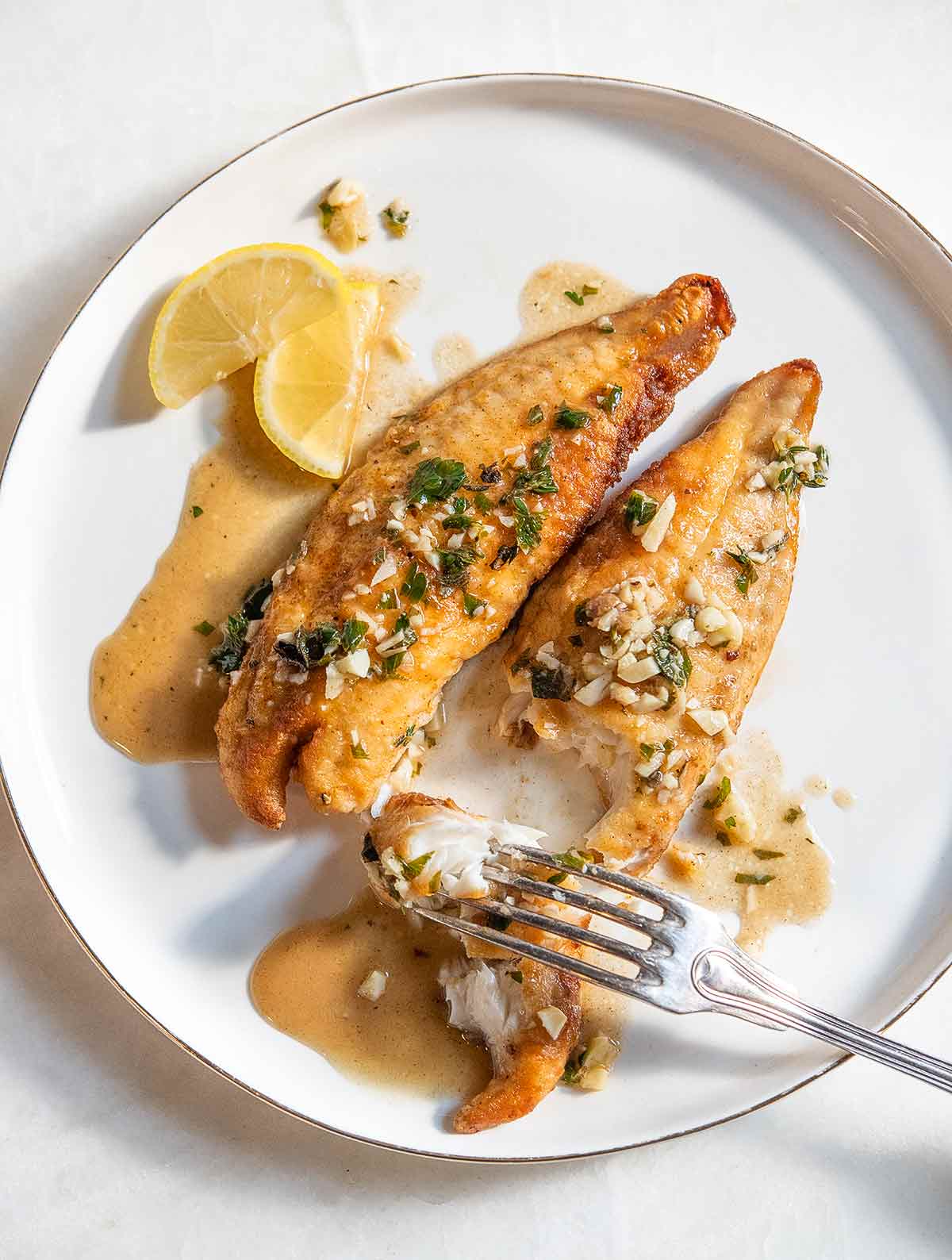 A white dinner plate with 2 filets of pan-fried bass with lemon garlic herb sauce, a fork, and 2 slices of lemon.