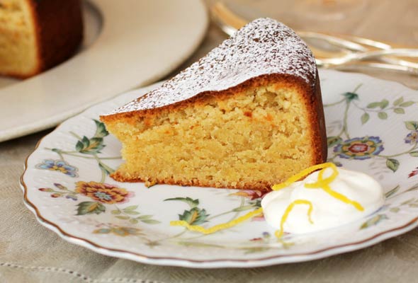 A floral plate with a slice of Portuguese almond torte, dusted with icing sugar, with a dollop of whipped cream and shreds of lemon zest.