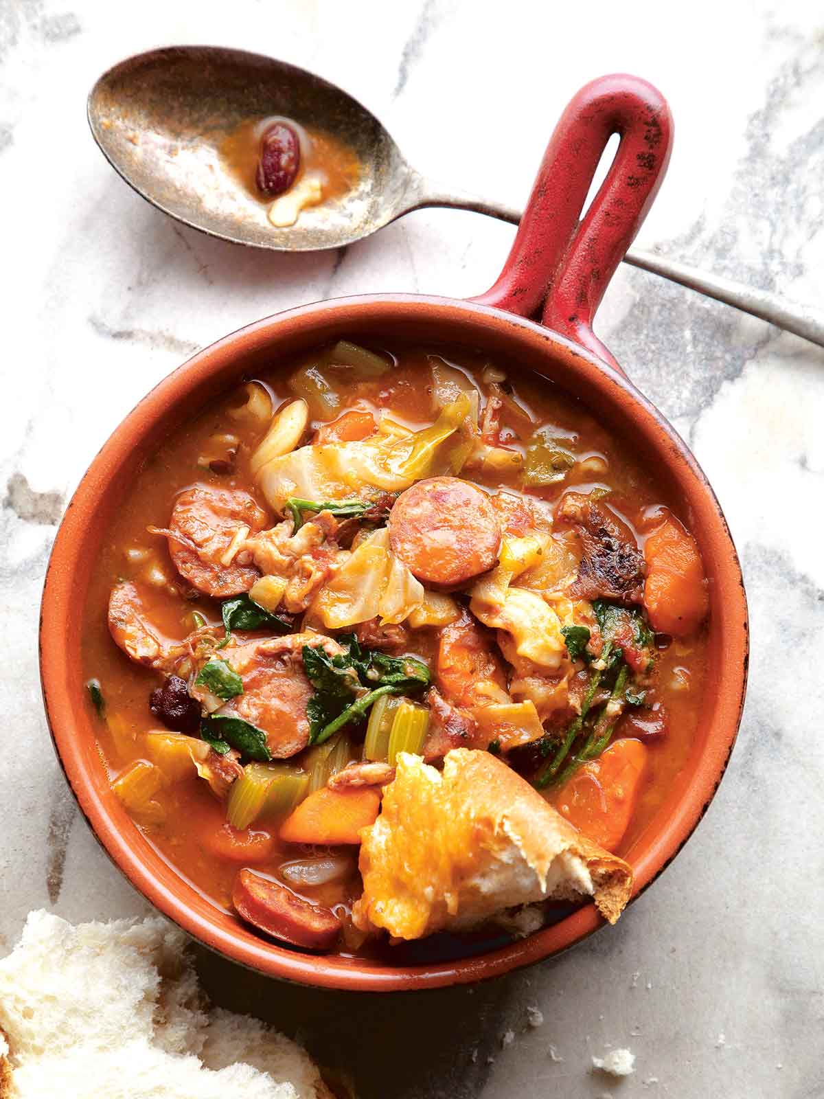 An enamel pot filled with Portuguese bean soup with a hint of bread roll and a spoon.