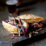 A wooden cutting board with two pulled pork sandwiches in crusty buns and topped with coleslaw, with a bottle and glass of beer in the background.
