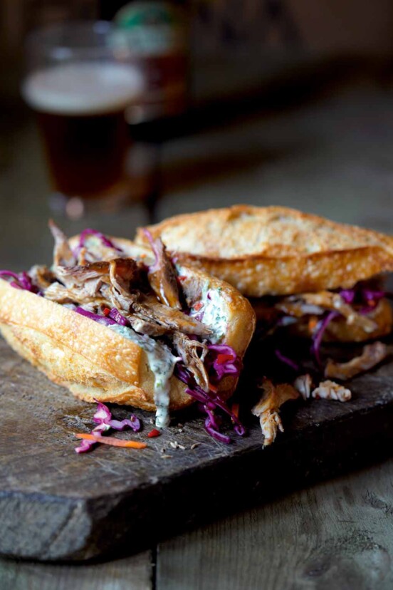 A wooden cutting board with two pulled pork sandwiches in crusty buns and topped with coleslaw, with a bottle and glass of beer in the background.