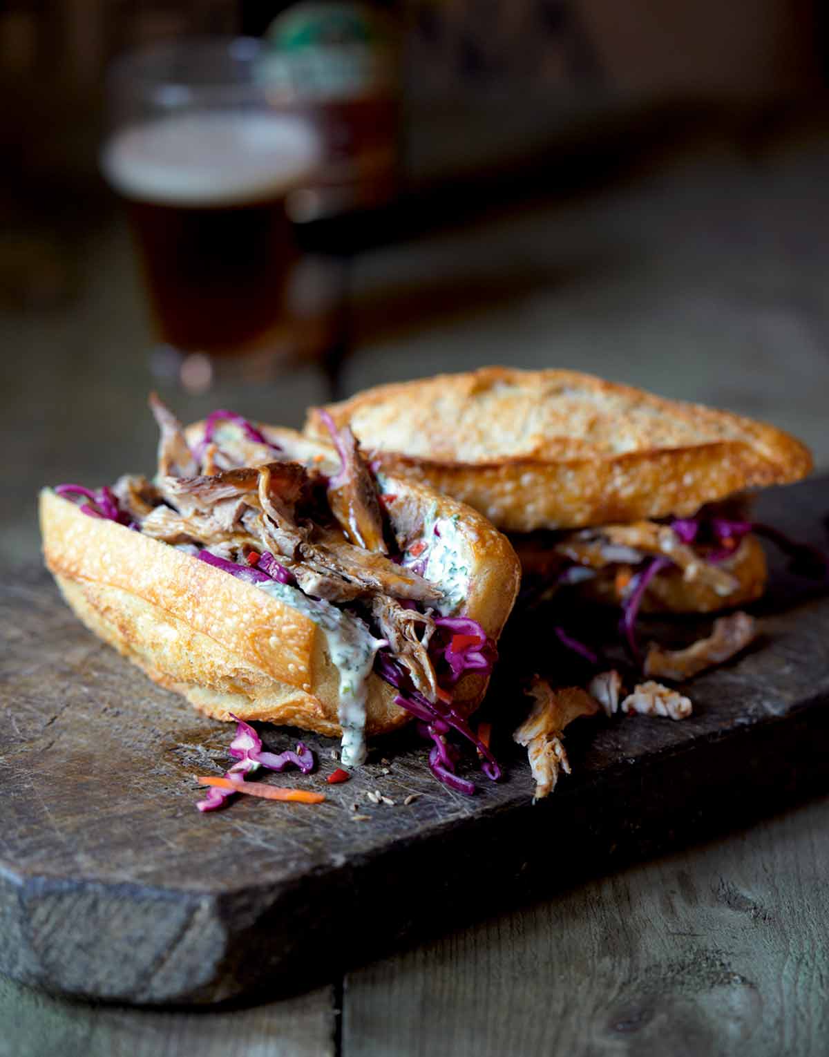 A wooden cutting board with two pulled pork sandwiches in crusty buns and topped with coleslaw, with a bottle and glass of beer in the background.