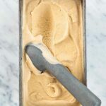 A metal container filled with pumpkin ice cream and a metal ice cream scoop resting inside.