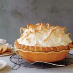 A pumpkin meringue pie in an orange stoneware pie plate, sitting on a cooling rack with a pie server.