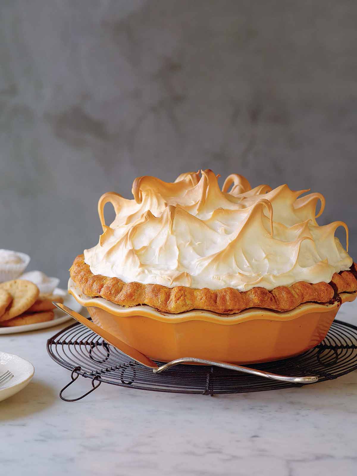 A pumpkin meringue pie in an orange stoneware pie plate, sitting on a cooling rack with a pie server.