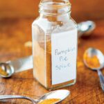 Pumpkin pie spice in a glass jar on a wooden cutting board, surrounded by measuring spoons full of various spices.
