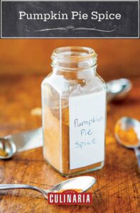 Pumpkin pie spice in a glass jar on a wooden cutting board, surrounded by measuring spoons full of various spices.