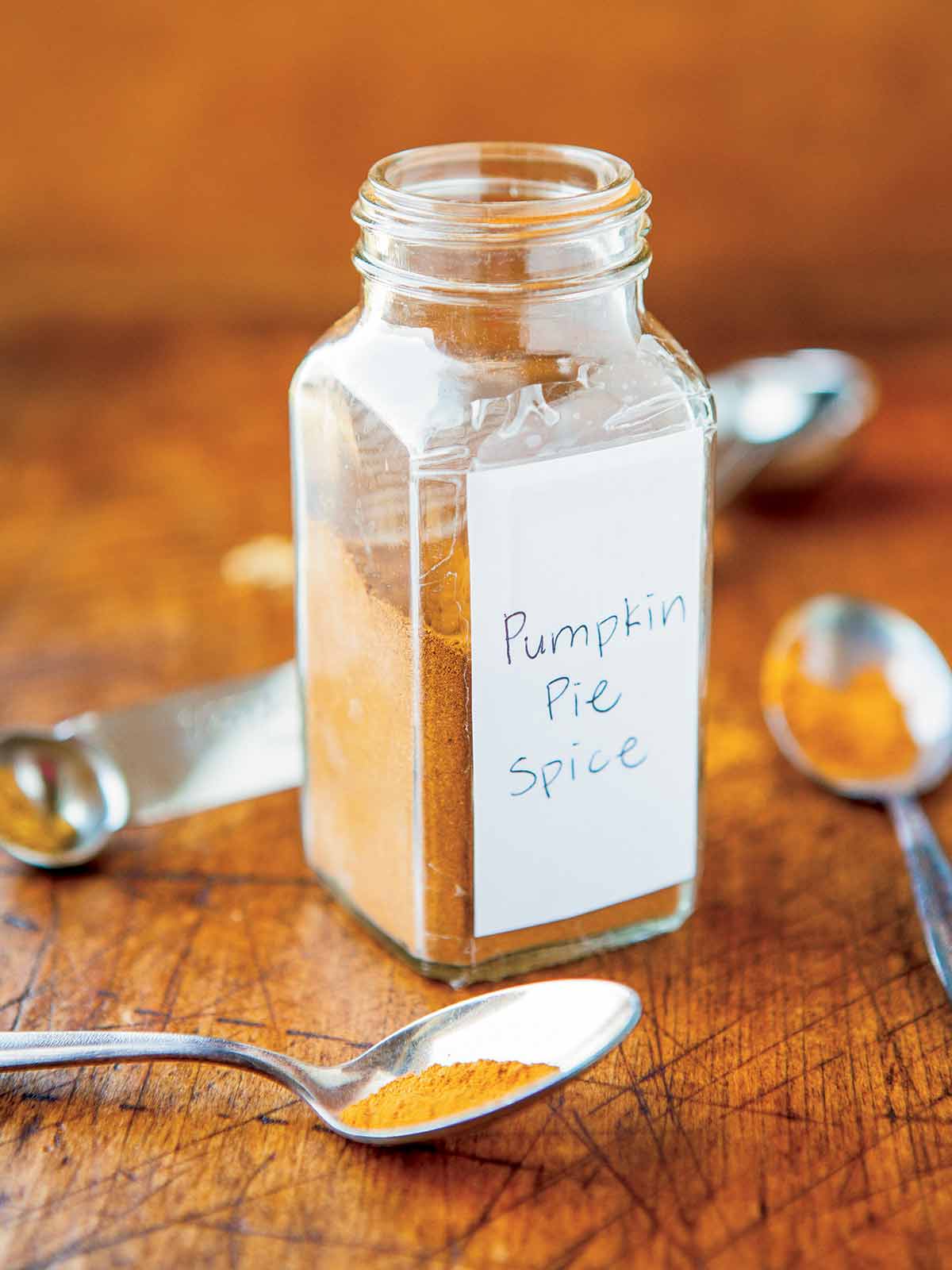 Pumpkin pie spice in a glass jar on a wooden cutting board, surrounded by measuring spoons full of various spices.